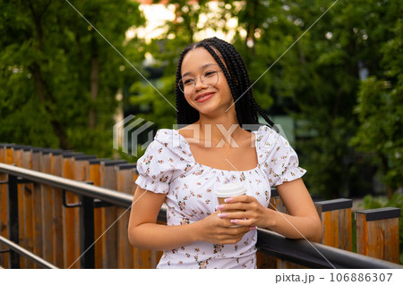 African young attractive woman in glasses stands on a bridge with a paper cup of coffee on the street during the day in the park in summer 106886307