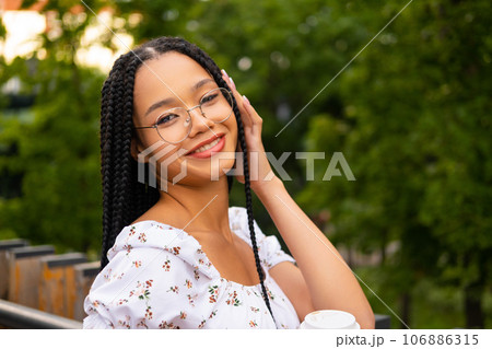 African young attractive woman in glasses stands on a bridge with a paper cup of coffee on the street during the day in the park in summer and looks at the camera African young attractive woman in glasses stands on a bridge with a paper cup of coffee on the street during the day in the park in summer and looks at the camera 106886315
