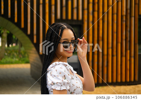 Young African beautiful woman holding dark sunglasses with her fingers smiling and looking at the camera on the street against the background of a wooden wall in the park Young African beautiful woman holding dark sunglasses with her fingers smiling and looking at the camera on the street against the background of a wooden wall in the park 106886345