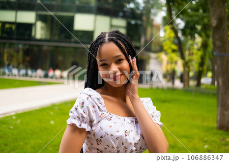 Portrait of a young pretty afro woman looking at the camera and smiling alone in the summer in the park. Touches face with beautiful manicured fingers Portrait of a young pretty afro woman looking at the camera and smiling alone in the summer in the park. Touches face with beautiful manicured fingers 106886347