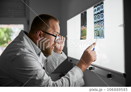 Side view of serious doctor studying MRI results. Male specialist working in hospital, carrying out description of radiographs of patient, pointing by pen. Concept of medicine and hospital. 106889585