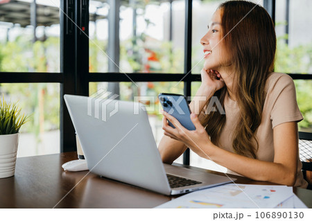 Happy business woman working with laptop computer she holding smart mobile phone at cefa for texting messages, lifestyle young female smiling using smartphone in coffee shop 106890130