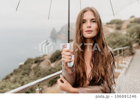 Woman rain umbrella. Happy woman portrait wearing a raincoat with transparent umbrella outdoors on rainy day in park near sea. Girl on the nature on rainy overcast day. 106891246