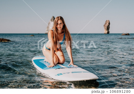 Woman sea sup. Close up portrait of happy young caucasian woman with long hair looking at camera and smiling. Cute woman portrait in a blue bikini posing on sup board in the sea Woman sea sup. Close up portrait of happy young caucasian woman with long hair looking at camera and smiling. Cute woman portrait in a blue bikini posing on sup board in the sea 106891280