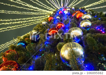 Multi-colored glass shiny balls on the New Year's city tree. Glowing garlands hang over the Christmas tree. Outdoor winter decoration. Festive design of the city square. Red, blue and yellow ball 106895082