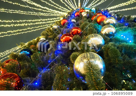 Multi-colored glass shiny balls on the New Year's city tree. Glowing garlands hang over the Christmas tree. Outdoor winter decoration. Festive design of the city square. Red, blue and yellow ball Multi-colored glass shiny balls on the New Year's city tree. Glowing garlands hang over the Christmas tree. Outdoor winter decoration. Festive design of the city square. Red, blue and yellow ball 106895089
