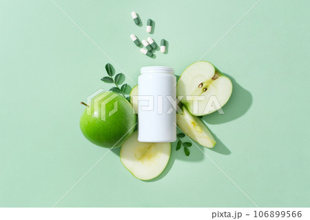 On the pastel background, a white medicine bottle unlabeled with capsule pills decorated with fresh slices of green apple. Advertising photo for product with natural extract for weight loss 106899566