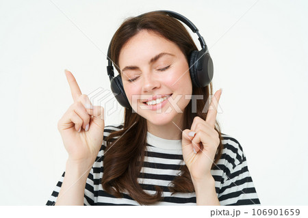 Portrait of happy student girl, listening music in headphones, dancing and pointing fingers at copy space, promo text, isolated against white background Portrait of happy student girl, listening music in headphones, dancing and pointing fingers at copy space, promo text, isolated against white background 106901659