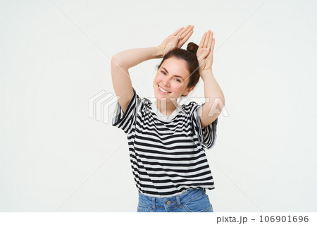 Cute young woman shows animal ears gesture above head, smiling and looking happy, posing in casual clothes over white background 106901696