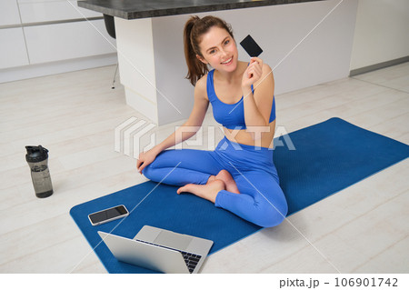 Portrait of young fitness girl paying for online class subscription, workout app, holding credit card, sitting on yoga mat with laptop, buying fitness tutorials 106901742