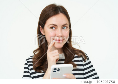 Portrait of smiling woman thinks to order something online on smartphone app, holding mobile phone with thoughtful face 106901758