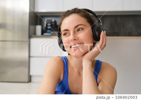 Close up portrait of brunette fitness girl, listens music in wireless headphones, smiles at camera, does workout alone at home, sits on floor and exercises 106902003