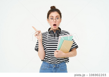 Portrait of girl gasps in surprised, points left with amazed face. Shocked student shows something, pointing finger at banner, holding notebooks, white background 106902203