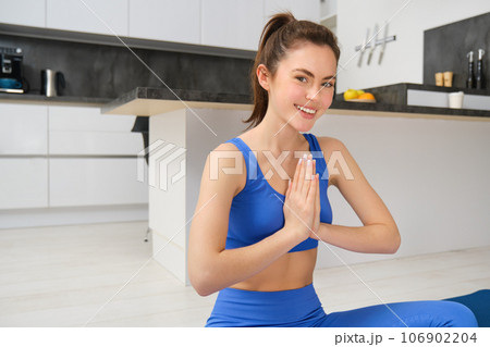Young woman, fitness trainer does yoga from home, clasps hands together, namaste gesture, sits with crossed legs on rubber mat, meditating from home and smiling 106902204