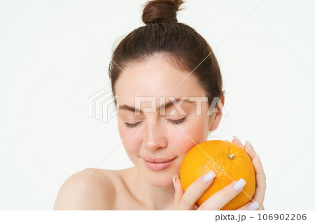 Close up portrait of brunette female model, holding orange near face, standing over white background 106902206