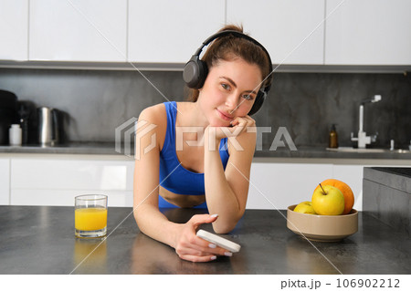Portrait of young fitness woman with headphones, drinking orange juice in kitchen and using smartphone, listening music, getting ready for workout gym Portrait of young fitness woman with headphones, drinking orange juice in kitchen and using smartphone, listening music, getting ready for workout gym 106902212