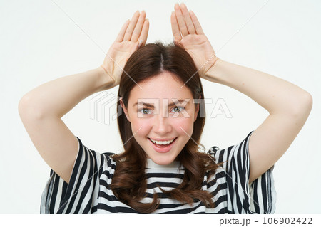 Image of cute brunette girl shows animal eats with hands on top of head, smiling and looking happy, white background 106902422