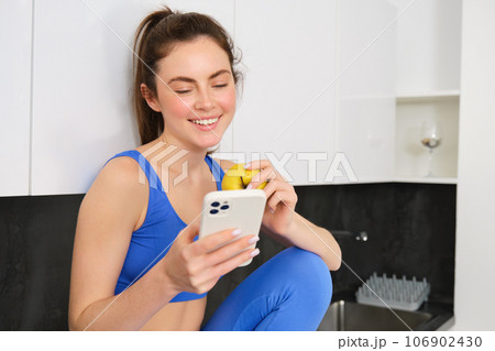 Healthy lifestyle and technology. Young beautiful girl, fitness instructor, looking at smartphone, reading on mobile phone and eating an apple, smiling happily 106902430