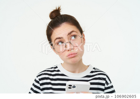 Close up portrait of girl in glasses, looks puzzled, holds phone and thinks, makes choice, decides what to order, does online shopping in mobile app, white background 106902605