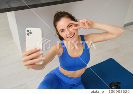 Portrait of young styling fitness girl doing workout from home, taking selfie and video for social media, gym instructor records her training session indoors 106902626