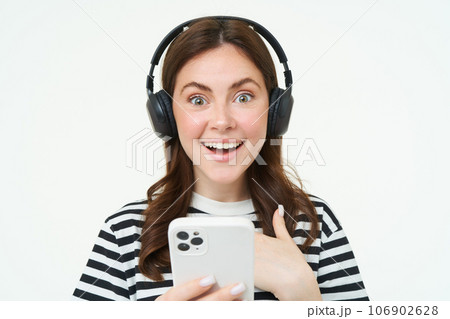 Young woman with surprised face, looking at smartphone screen, reacts amazed, looks with happiness at mobile phone, standing in wireless headphones, white background Young woman with surprised face, looking at smartphone screen, reacts amazed, looks with happiness at mobile phone, standing in wireless headphones, white background 106902628