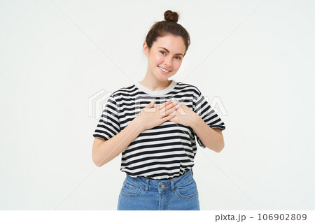 Young smiling woman, feels gratitude, thankful for something, holds hands on heart and looks tender at camera, standing over white background 106902809
