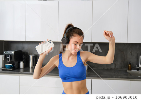 Portrait of beautiful fitness blogger, woman in headphones, listening music and dancing in kitchen, wearing blue leggings and sportsbra 106903010
