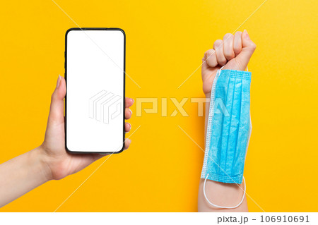 One woman's hand is holding a medical mask, while the other hand is holding a cellphone. Mock up. Yellow background. Concept of protection against coronavirus and other diseases 106910691