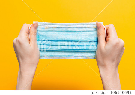 Female's hands hold tight medical mask. Yellow background. Close up. Concept of protection against coronavirus and other diseases Female's hands hold tight medical mask. Yellow background. Close up. Concept of protection against coronavirus and other diseases 106910693
