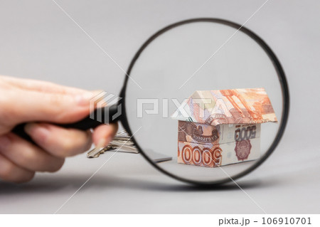 Female hand holding a magnifier glass and showing zoomed at paper origami from russian rubles. Gray background. Concept of mortgage and leasing of real estate 106910701