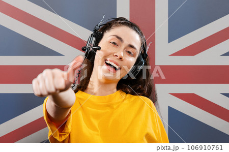 World English Language Day. Portrait of a young smiling woman wearing headphones, giving a thumbs-up. On the background of the English flag. The concept of learning foreign languages 106910761