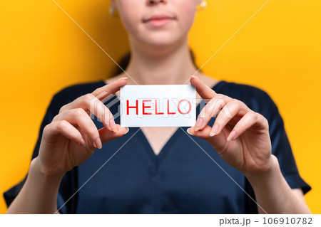 World Hello day. A close-up portrait of a woman holding a white business card with the inscription Hello. Yellow background World Hello day. A close-up portrait of a woman holding a white business card with the inscription Hello. Yellow background 106910782