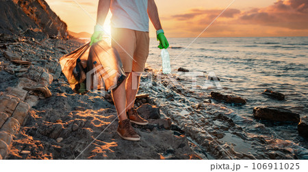 A male volunteer holding two polyethilene bag with garbage. In the background the sea and the sunset. Banner with copy space. The concept of environmental protection and coastal clean up 106911025