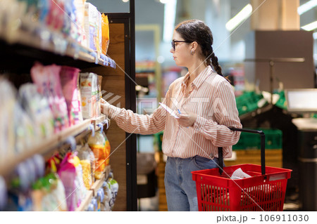 Sale in grocery store. Side view of young caucasian woman wearing eyeglasses holds cart and take food from upper shelf. Concept of shopping in supermarket and consumerism Sale in grocery store. Side view of young caucasian woman wearing eyeglasses holds cart and take food from upper shelf. Concept of shopping in supermarket and consumerism 106911030