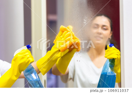Woman in rubber gloves sprays cleanser and wipes mirror with rag. View from shoulder. Cleaning service and chores 106911037
