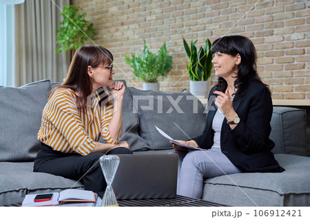 Two business mature women sitting on couch in office, talking, discussing work Two business mature women sitting on couch in office, talking, discussing work 106912421