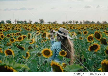 Woman in Sunflower Field: Happy girl in a straw hat and blue dress stands in a vast field of sunflowers at sunset. Summer time. 106917704