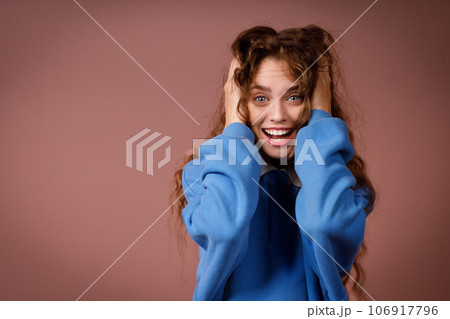 Happy surprised young adult woman wearing winter blue sweater and hold head with her hands looking at camera with joyful smile Happy surprised young adult woman wearing winter blue sweater and hold head with her hands looking at camera with joyful smile 106917796
