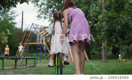 Young mother holds bicycle while little daughter learns to ride on playground Young mother holds bicycle while little daughter learns to ride on playground 106918480