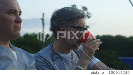 Two doctors sit on bench, drink coffee during break, discuss work Two doctors sit on bench, drink coffee during break, discuss work 106919380