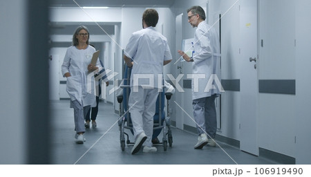 Doctors pushing transfer wheelchair with patient in clinic corridor 106919490