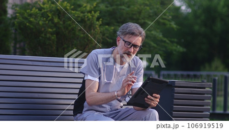 Doctor in uniform sits on bench, talks to patient using digital tablet Doctor in uniform sits on bench, talks to patient using digital tablet 106919519