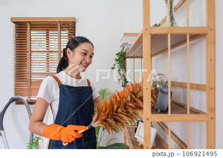 Asian woman cleaning in living room at home. Young woman housekeeper cleaner feel happy and use feather duster wiping messy dirty for housekeeping housework 106921495