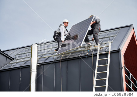 Installers installing solar panel system on roof of house. Men workers in helmets carrying photovoltaic solar module outdoors. Concept of alternative and renewable energy. Installers installing solar panel system on roof of house. Men workers in helmets carrying photovoltaic solar module outdoors. Concept of alternative and renewable energy. 106929984