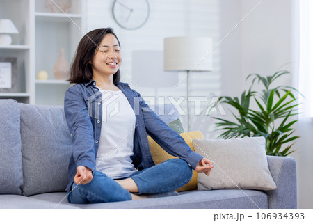 Young smiling asian woman meditating sitting on sofa in living room, woman smiling joyfully with eyes closed, dreaming and thinking about future plans, visualizing achievement. 106934393