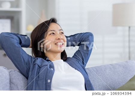 Closeup photo of a young beautiful Asian at home in the living room, woman has her hands behind her head and is resting, breathing fresh air, smiling and looking window, dreaming happy future. Closeup photo of a young beautiful Asian at home in the living room, woman has her hands behind her head and is resting, breathing fresh air, smiling and looking window, dreaming happy future. 106934394