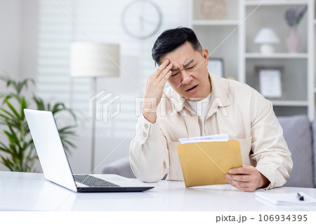 Upset and disappointed man in home office received message mail envelope with bad news, asian man working in home office at desk with laptop. Upset and disappointed man in home office received message mail envelope with bad news, asian man working in home office at desk with laptop. 106934395