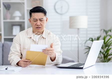 Upset and disappointed man in home office received message mail envelope with bad news, asian man working in home office at desk with laptop. 106934396