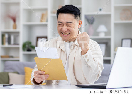 Happy and successful asian received mail notification envelope with good news, man sitting at table at home, working in home office in living room, joyfully celebrating winning news. 106934397