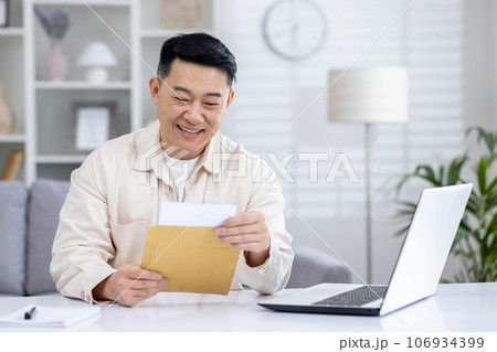 Happy and successful asian received mail notification envelope with good news, man sitting at table at home, working in home office in living room, joyfully celebrating winning news. 106934399